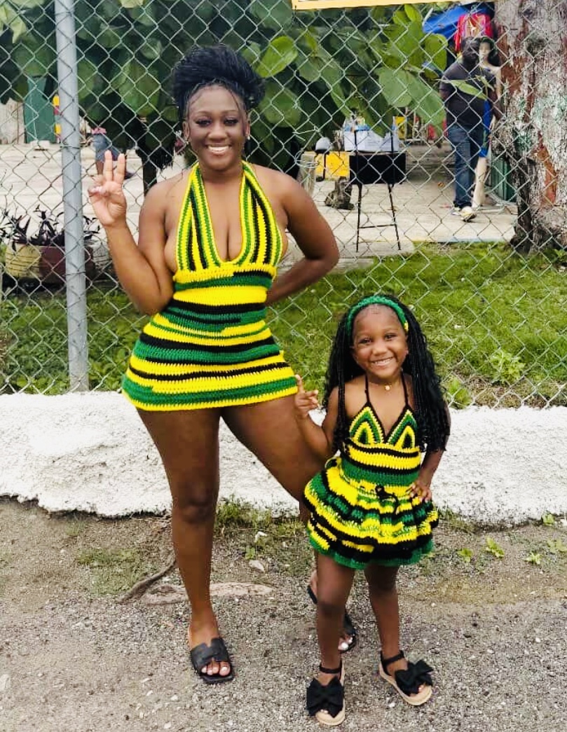 Two smiling females, one adult and one child, wearing matching yellow, green, and black striped dresses, standing outdoors near a fence.