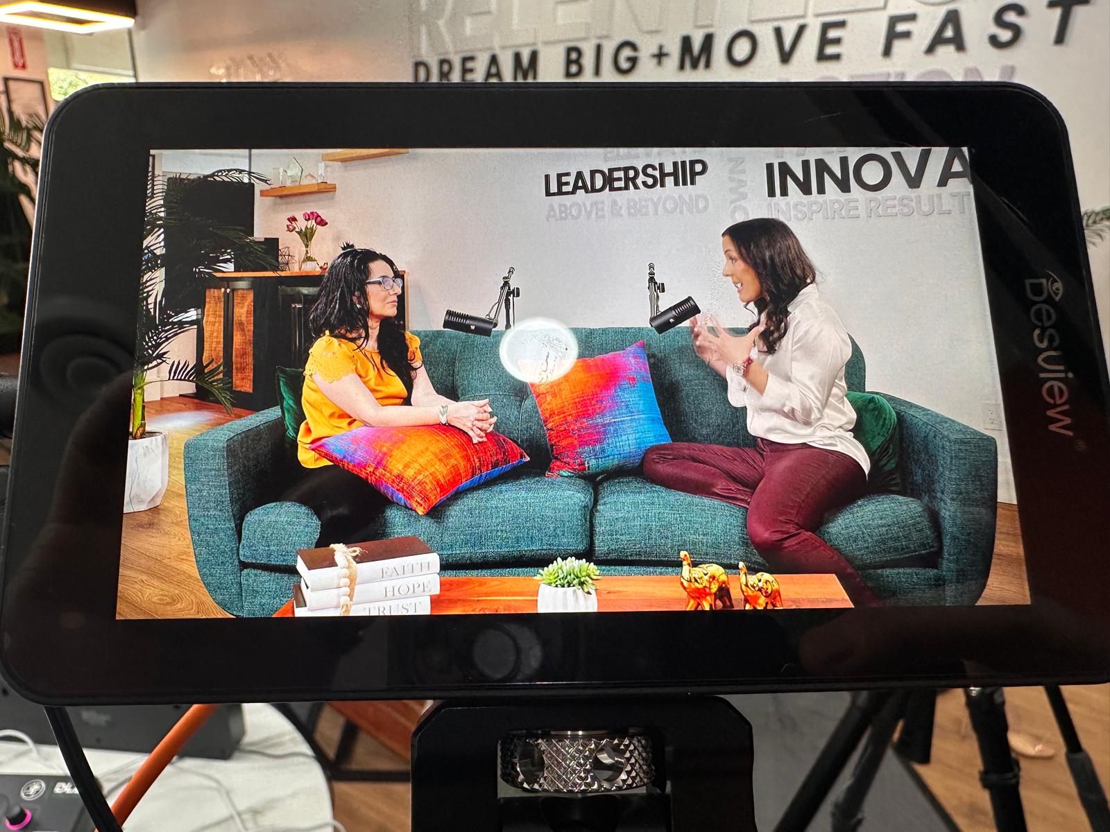 Two women sit on a sofa with colorful pillows, talking and reading a book in a modern room.