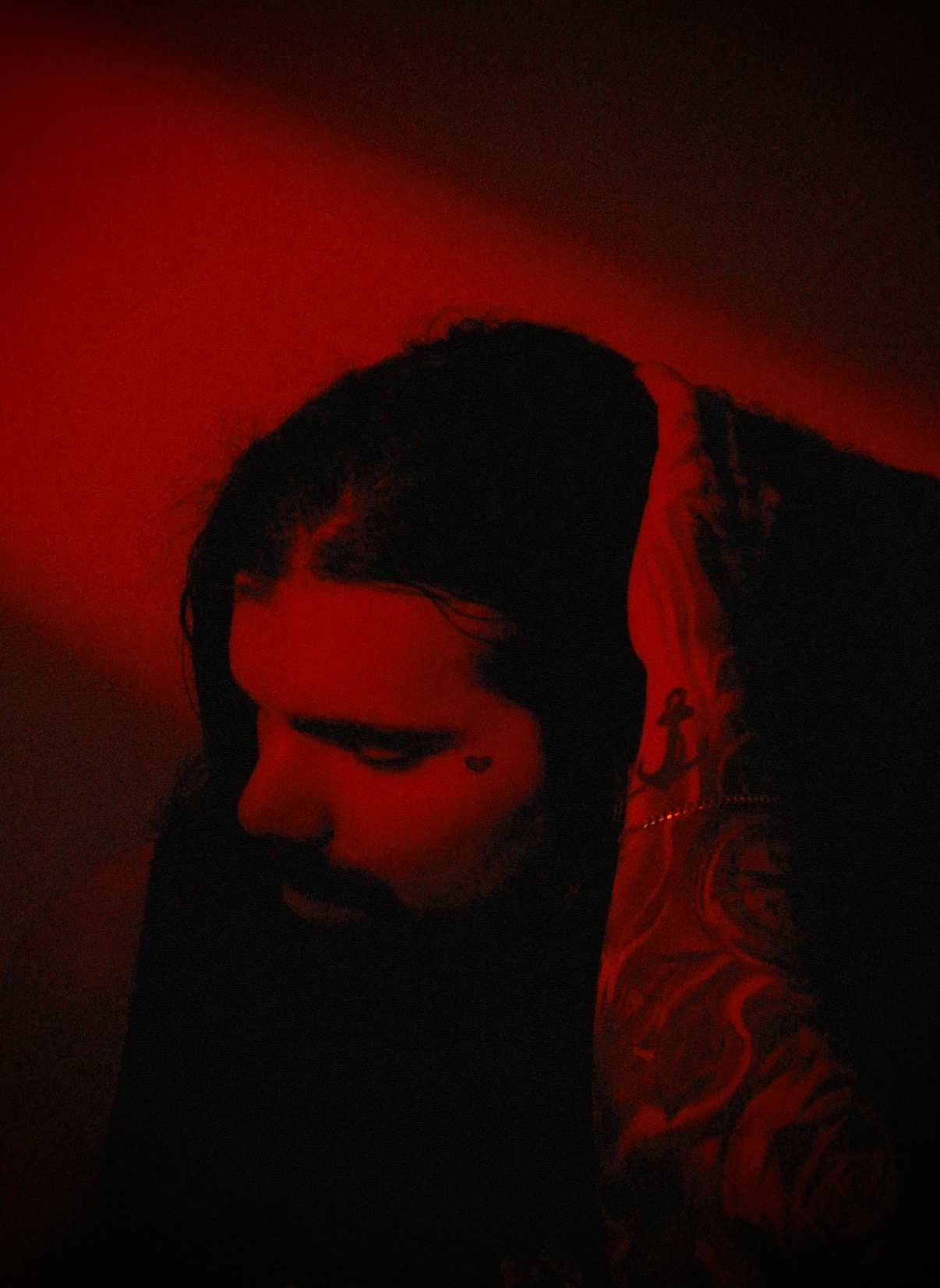 Person with long dark hair and a tattoo on their arm, illuminated by red light, looking downward.