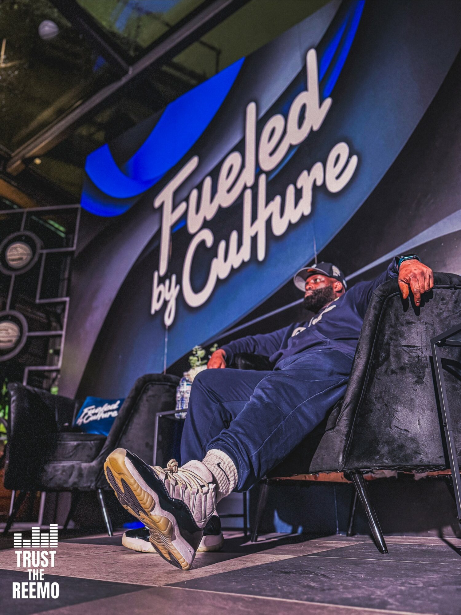 Person sitting on a stage with a large screen behind displaying 'Fueled by Culture' in white text, wearing casual clothing and sneakers.
