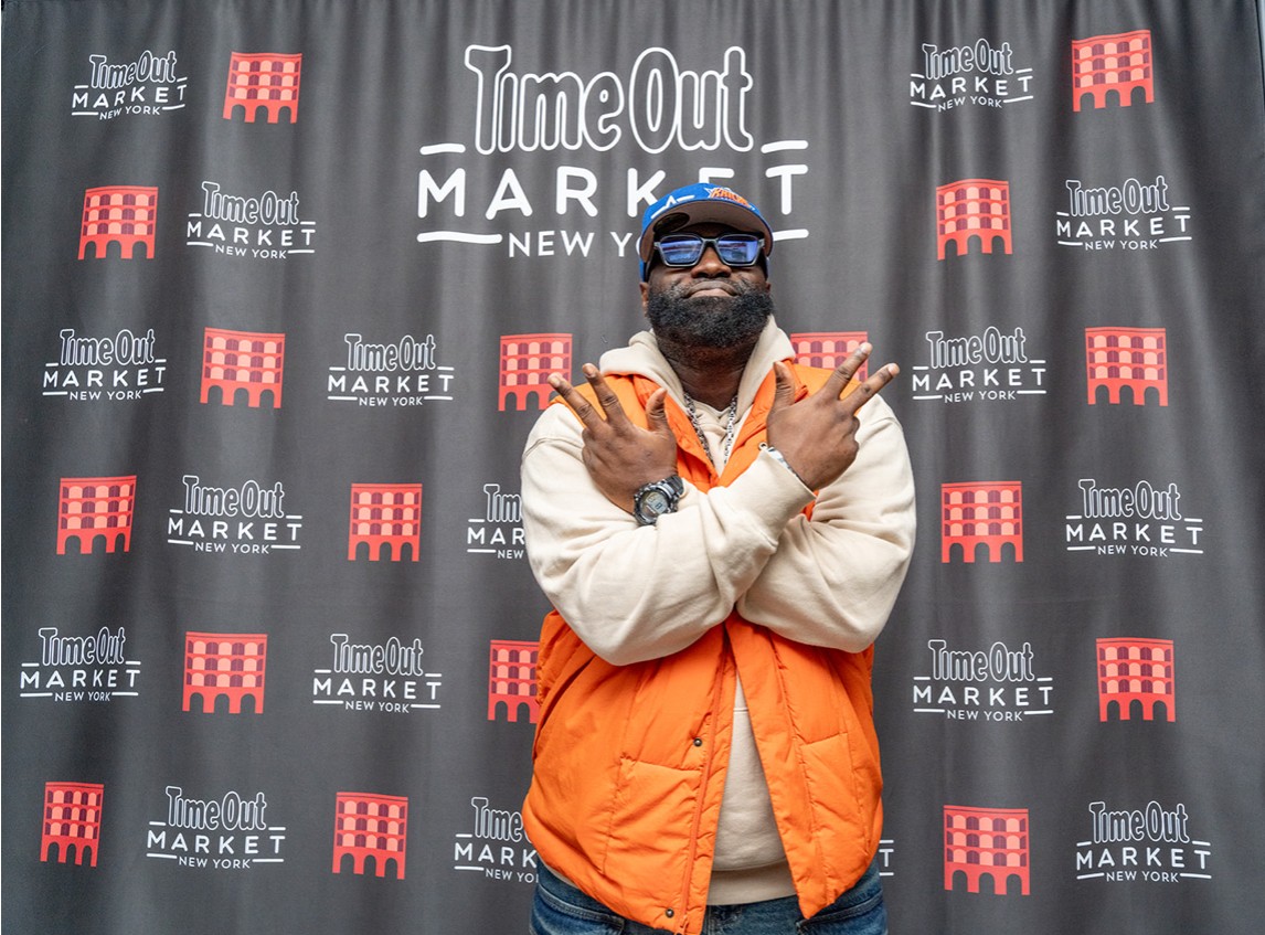 Man with sunglasses and beard wearing an orange and white jacket, crossing arms in front of a black backdrop with logos.