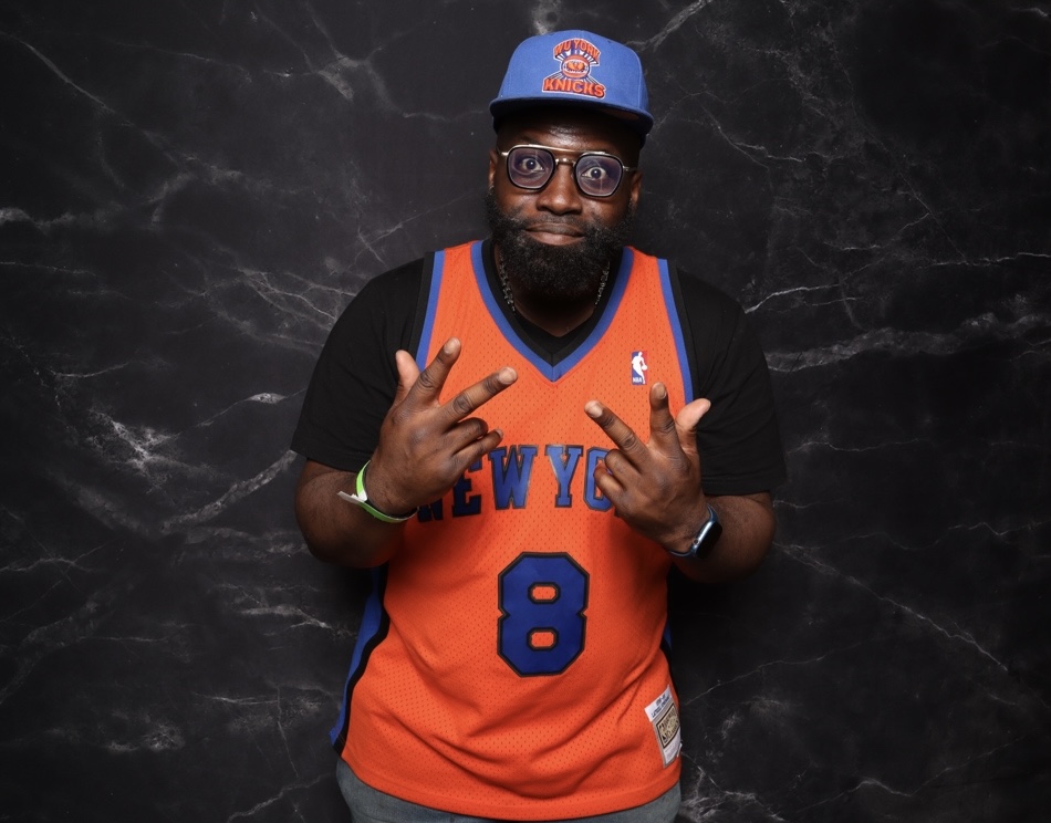 Person with glasses wearing a New York Knicks basketball jersey, cap, and making a peace sign gesture, standing against a dark background.