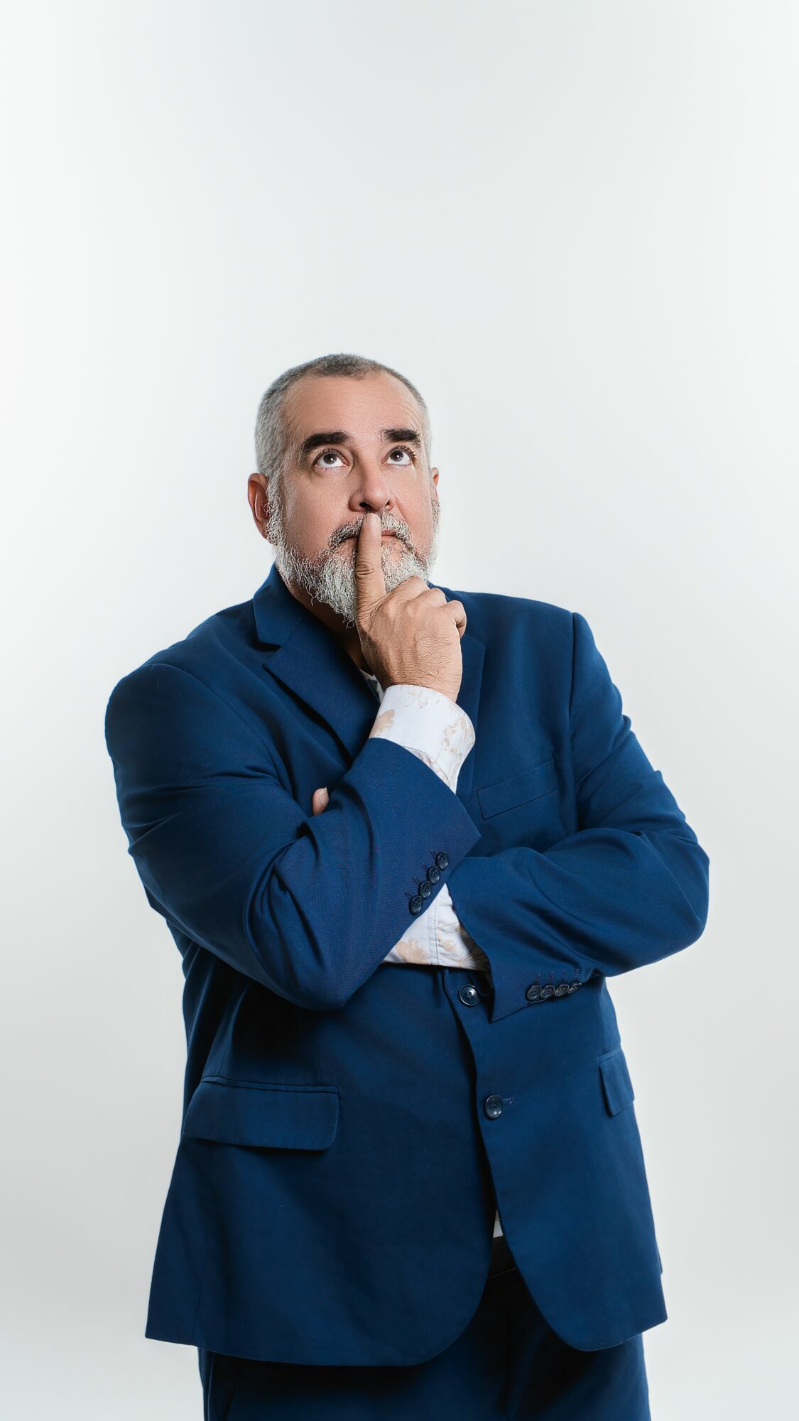 Man in a blue suit with hand on chin, looking upward, against a plain white background.