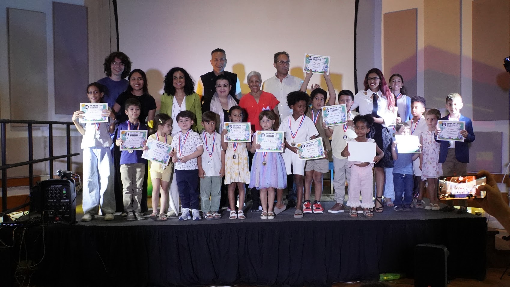 Group of children and adults on stage holding certificates, some taking photos, in a celebration setting.