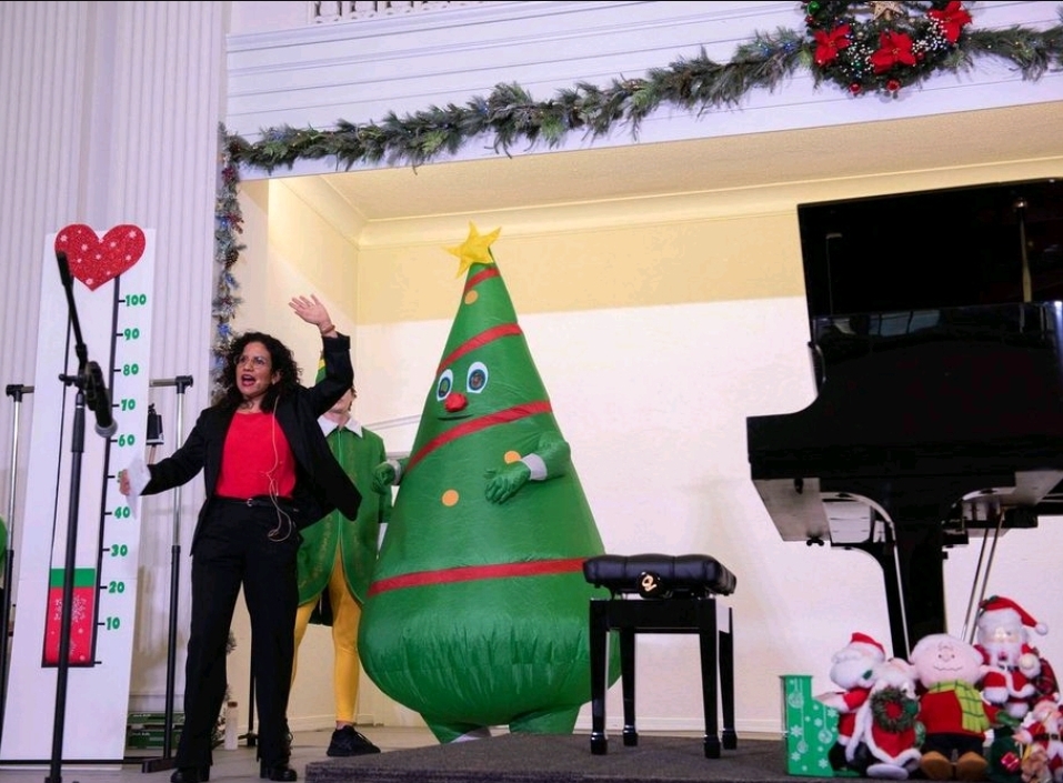 Woman standing on stage with Christmas decorations, including a large inflatable Christmas tree, a piano, and holiday props.